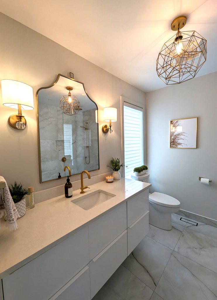 Bright vanity area with arched mirror, gold faucet, soft wall sconces, and marble-look flooring — luxury bathroom by Lucie Pitt.