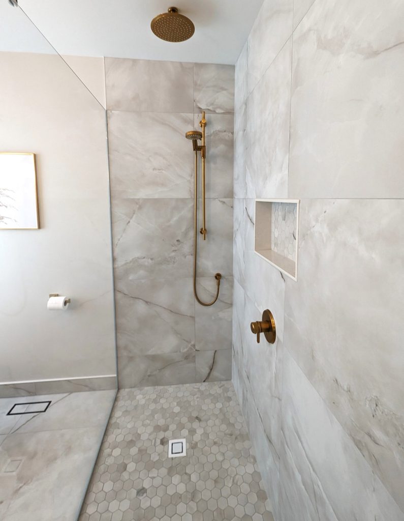 Elegant walk-in shower with brass rain showerhead, marble-look porcelain wall tiles, and hexagon mosaic flooring from the West Island Luxe project by Lucie Pitt.