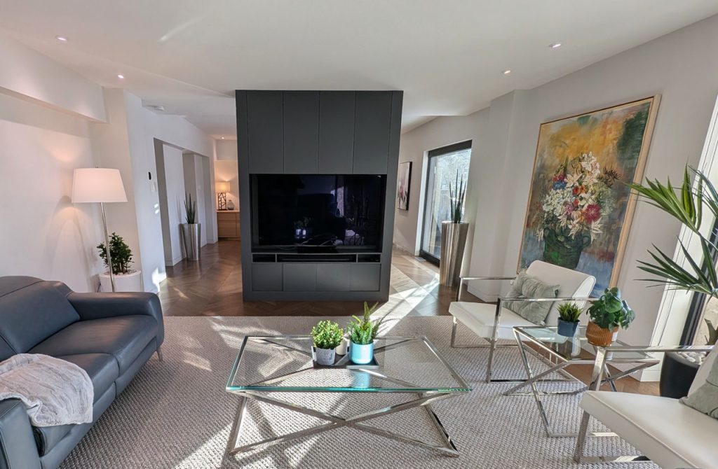 Modern living room with sleek charcoal built-in TV wall, glass tables, natural light, and contemporary seating — Habitat 67 refinement by Lucie Pitt.